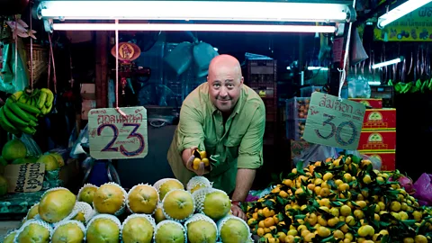 Andrew Zimmern bends over a fruit stand and holds small orange fruits in his hand (Credit: Courtesy of The Travel Channel)