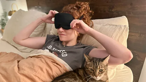 Ally Hirshlag, a woman with red curly hair wearing a black sleep mask, wakes up in bed with her cat beside her. She is pulling off her sleep mask (Credit: Ally Hirschlag)