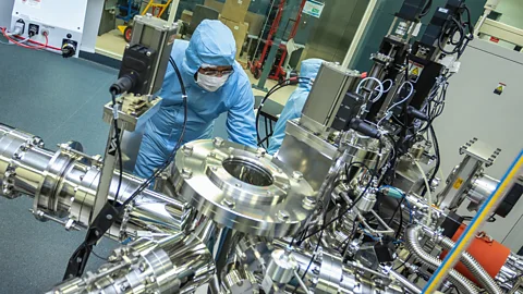 A man in a blue protective lab suit wearing a white face-mask leans over a large cylindrical metal machine in a lab (Credit: Csiro)