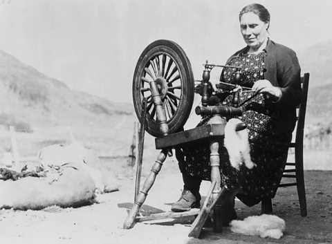 A middle-aged woman seated outside operating a spinning wheel