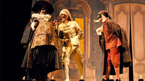 Three males stand on stage in highly-decorated costumes and masks that are indicative of commedia dell’arte.