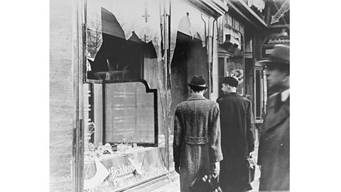 A black and white photograph of people walking past a window that has been smashed during ‘KٲԲ’.