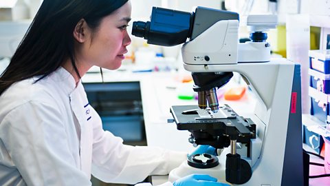 A scientist in a white lab coat and blue gloves looks through a large microscope.