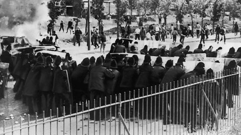 A photograph showing lines of police with riot shields face a group of youths during riots in the Toxteth area of Liverpool, July 1981.