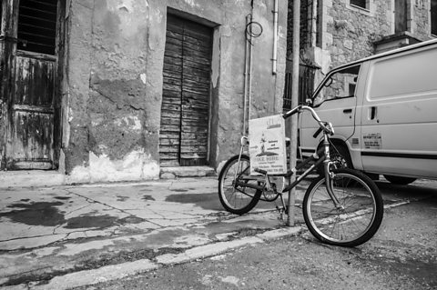 Black and white student street photography image of a bicycle leaning against a lamppost in front of buildings with flaking plaster and worn wooden shutters.