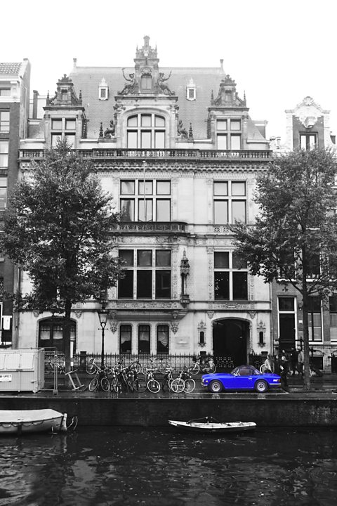 Black and white photograph of a grand house overlooking a river with a blue sports car in colour