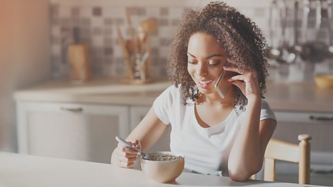 Woman eating breakfast in kitchen while talking on her smart phone