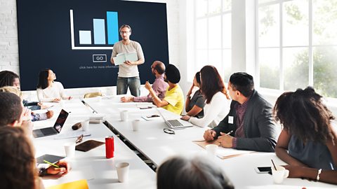 Man front of a large screen giving presentation to a group sitting at a large table