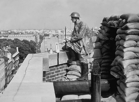 A man in military uniform and helmet stands watch with a bell and ratchet on top of a tall building overlooking a city.
