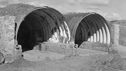 Two metal half-cylinders partially covered in earth. Crates can be seen in one of the structures.