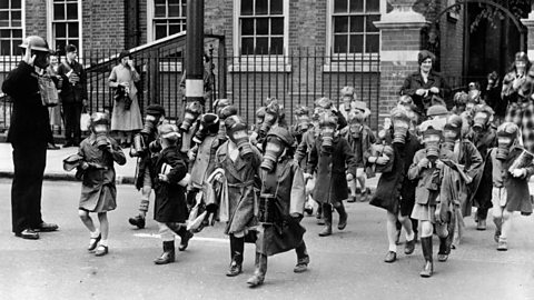 Young children walking in a group, all wearing gas masks. They are saluted by a man an air raid warden in a gas mask.
