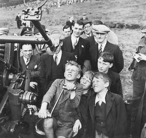 A group of men and boys look jovial and interested as they inspect Anti-aircraft gun on display to the public.
