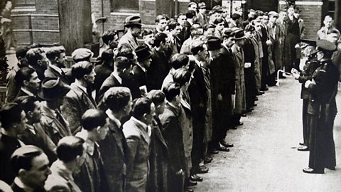 Men in WWII era clothing standing in a group forming lines. Men in uniform with a clipboard address them.