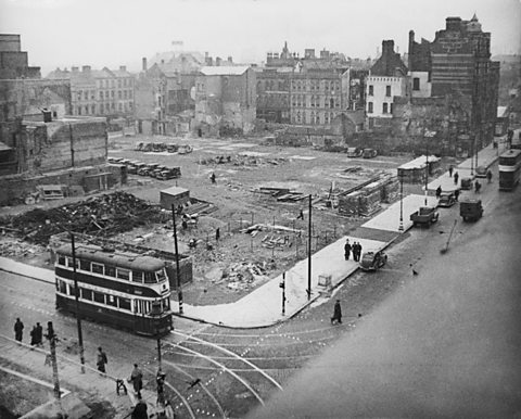 Golygfa o strydoedd Belfast gyda bloc sydd bron yn wag, a thomennydd o rwbel lle’r arferai adeilad fod.