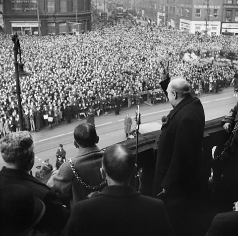 Winston Churchill yn sefyll ar falconi gerbron torf anferth yn rhoi’r arwydd ‘V for victory’ gyda’i law.