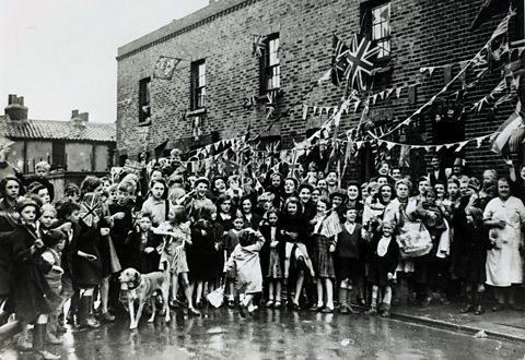 Large group of people smiling and waving in front of their terraced houses adorned with bunting and flags.