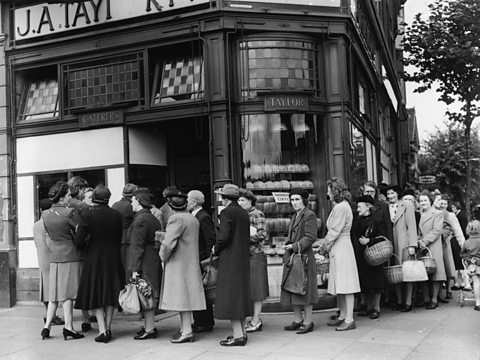 Women in 1940s clothing queue with baskets outside a bakery.