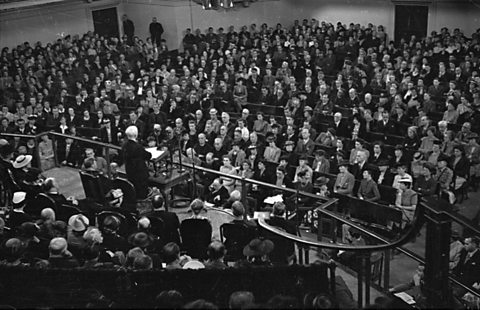 William Beveridge addresses a crowd from a podium in a hall.