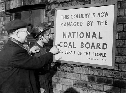 Two men, one in a mining helmet look at a poster that reads; This colliery is now managed by the National Coal Board on behalf of the people.