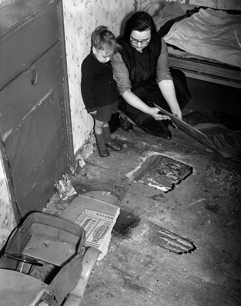 A woman patching a hole in the floor of a dilapidated room. Her young son looks on