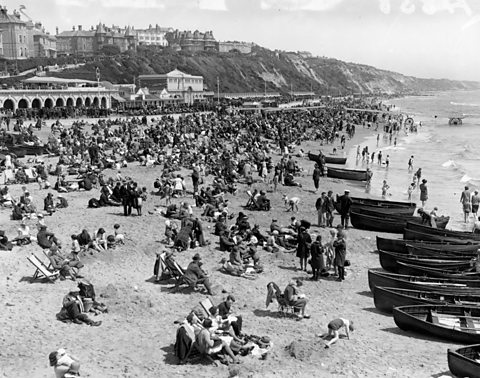 A crowded beach full of beachgoers. Building, cliffs and a promenade can be seen in the background. Boats wait on the shoreline.