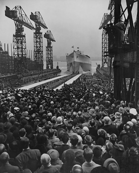 A massive crowd stand below large cranes in a dockyard, a HMS Prince of Wales can be seen in the background.
