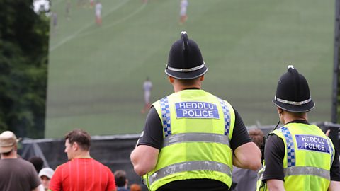 A photograph of two policemen in luminous jackets with their backs to the camera. People are watching a football match being shown on a large screen.