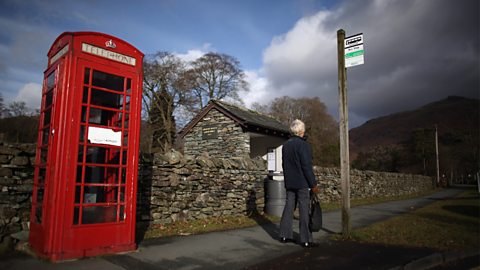 An elderly person waits at a rural bus stop. Next to the bus stop is a traditional red telephone box.