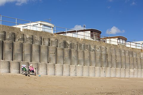 An old woman sitting in a deck chair on a beach in front of concrete sea defences. Behind them is a caravan park.