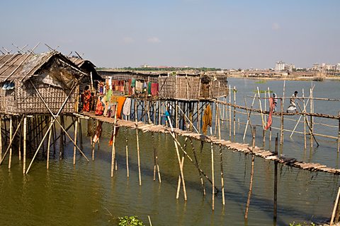 Traditional stilt houses standing in water, with high rise apartment buildings in the background. 