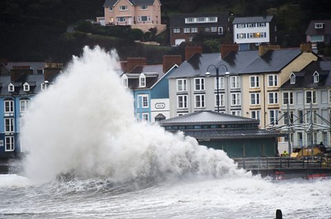 A large wave breaks on the seafront wall pushing white spray high into the air. 