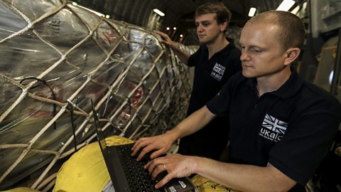 Aid items are held in large nets inside an aircraft. A man in the foreground is typing on a laptop computer. 