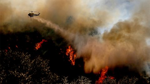 A helicopter is flying through clouds of smoke dropping water on a burning forest below.