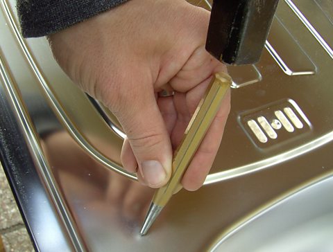 Hand holds centre punch above metal sheet which is about to be struck with a hammer