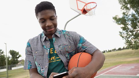 Teenage boy with basketball messaging on his mobile phone on basketball court.