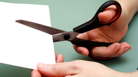 A pair of hands shown holding a piece of white card and a pair of scissors cutting it.
