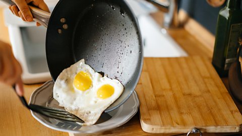 Some hands moving two fried eggs with a spatula from a black non-stick pan to a plate.