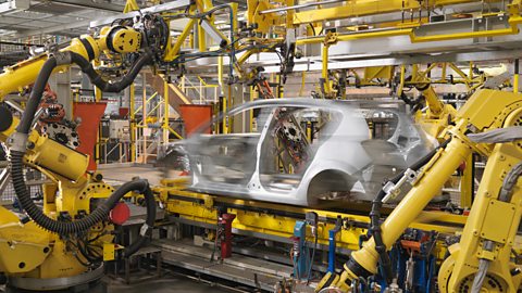 Multiple yellow robot arms working on building a silver car in a manufacturing plant.