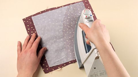 A pair of hands shown ironing a material onto a piece of burgundy polka dot fabric.