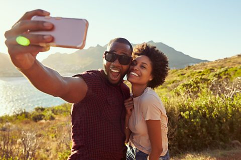 A couple are smiling and taking a selfie in the countryside in front of a lake.