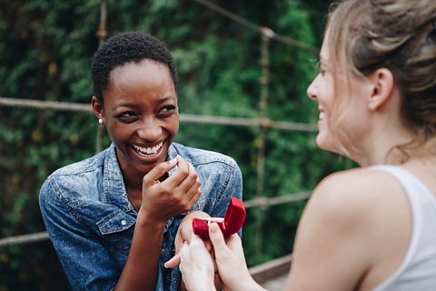A woman is proposing to her overjoyed girlfriend. There are trees in the background
