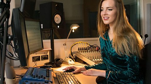 a young woman sits at a desk with computers and music composition technology