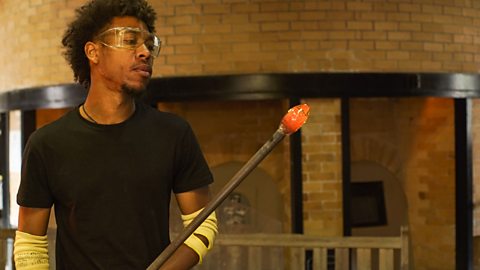 a young man stands holding a glass working tool, glowing red with heat