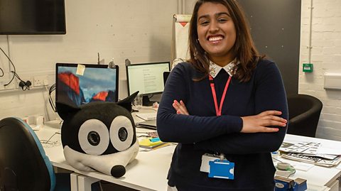 A young woman stands smiling at the camera in front of her busy desk, with her arms folded