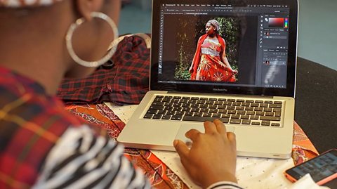 a young woman views photos of a fashion model, wearing the clothes she has designed on a laptop