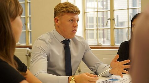 a young man sits a table, talking in a meeting