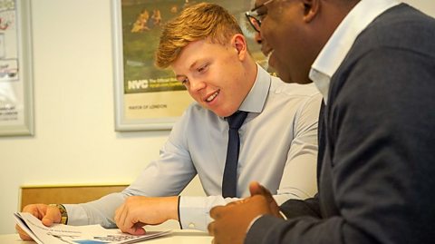 a young man reads documents at a table with a colleague