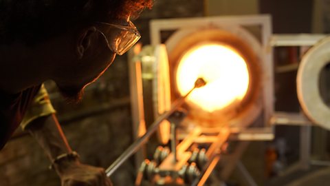 Jahday holds a long pole with molten glass into a fire