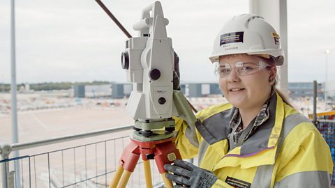 Sally wears a hard hat and operates a surveying machine on a construction site 