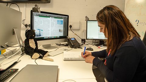 A young woman, Rosie, at her desk writing on a notepad with a couple of computers in front of her